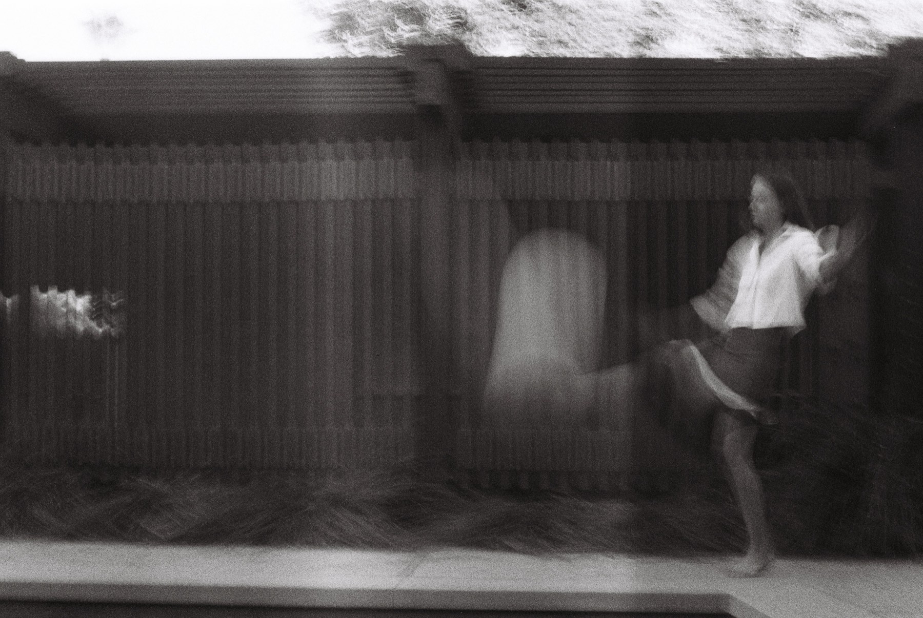 Black-and-white long-exposure photograph of a figure leaning against a slatted wooden wall, motion blur trailing into a second form.