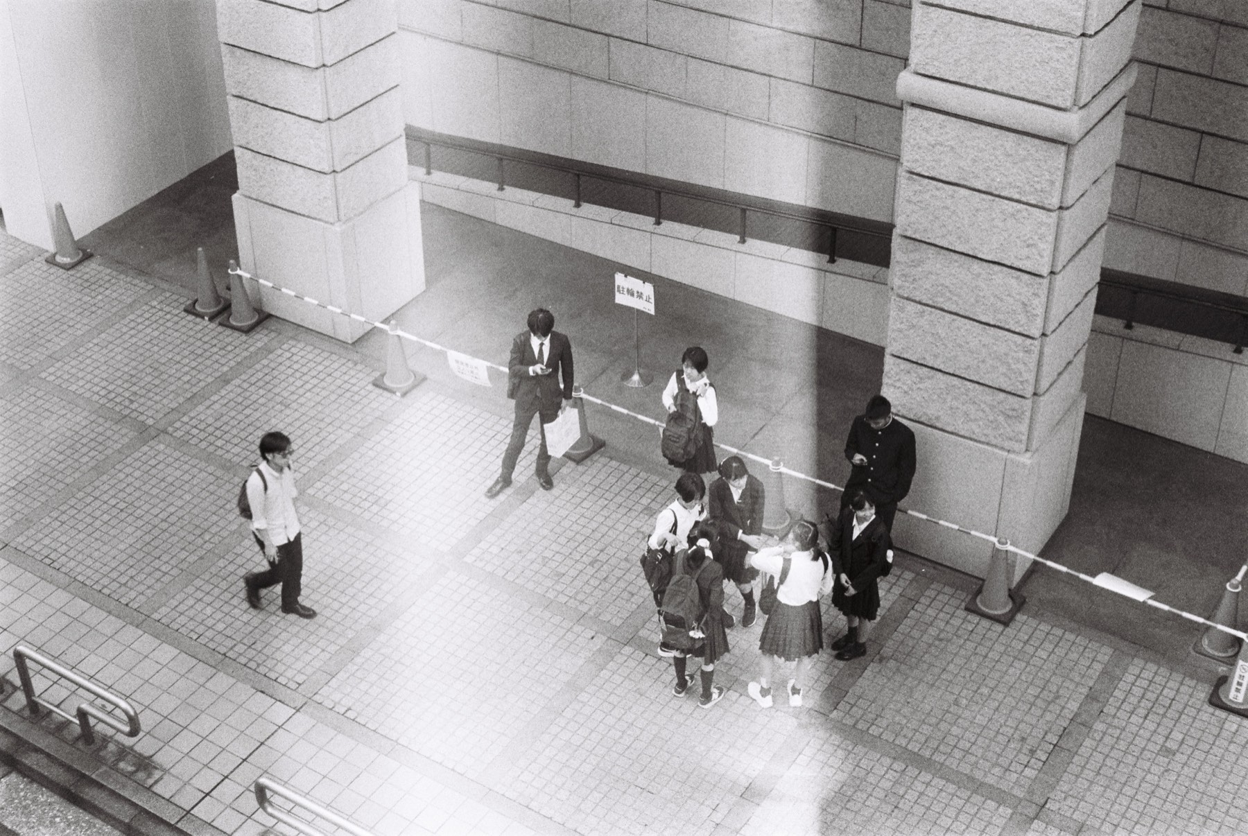 Black-and-white photograph, shot from above, of people gathered on a paved plaza between stone pillars.