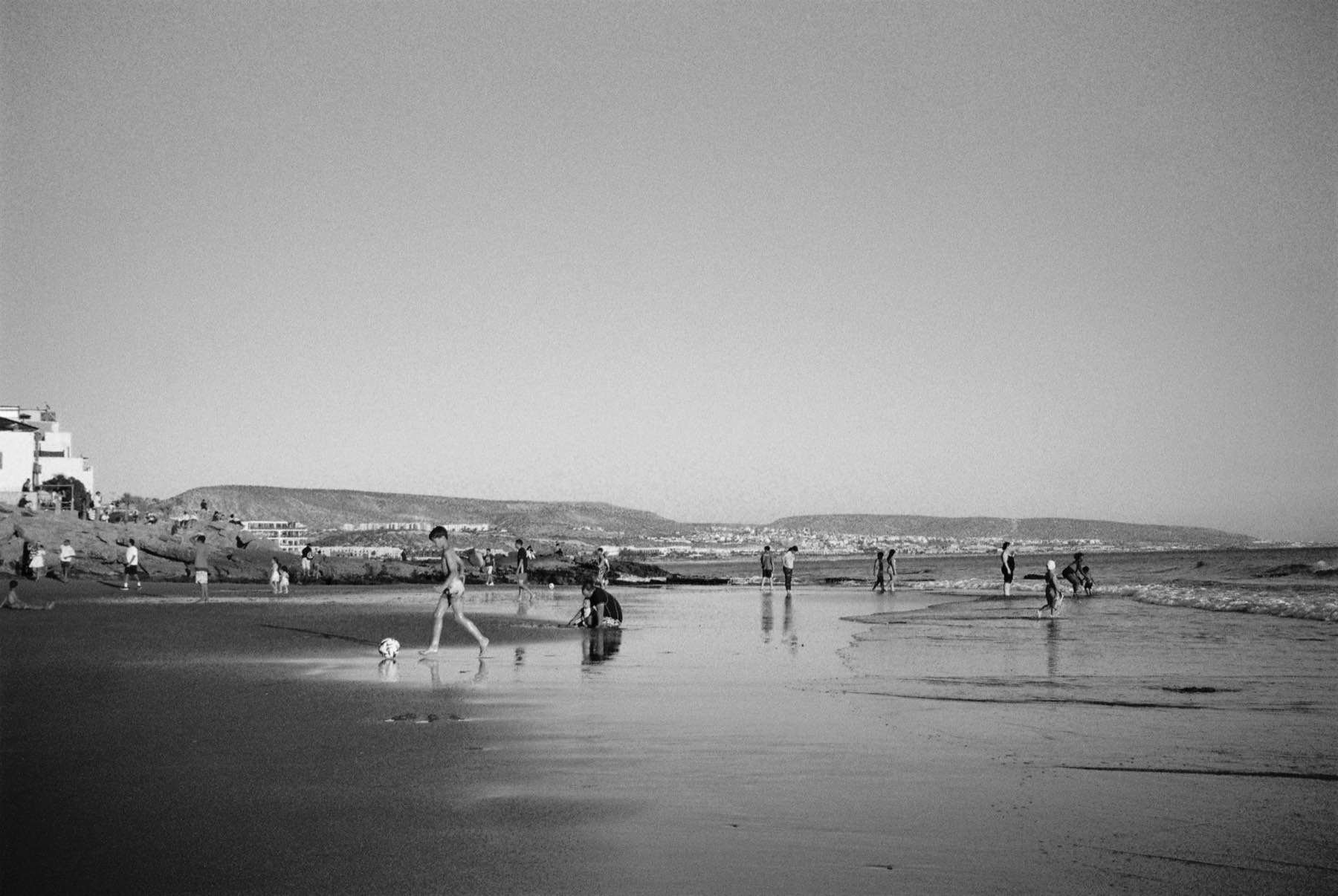 Black-and-white photograph of a wide beach at low tide, small figures and a dog on reflective sand beneath open sky.