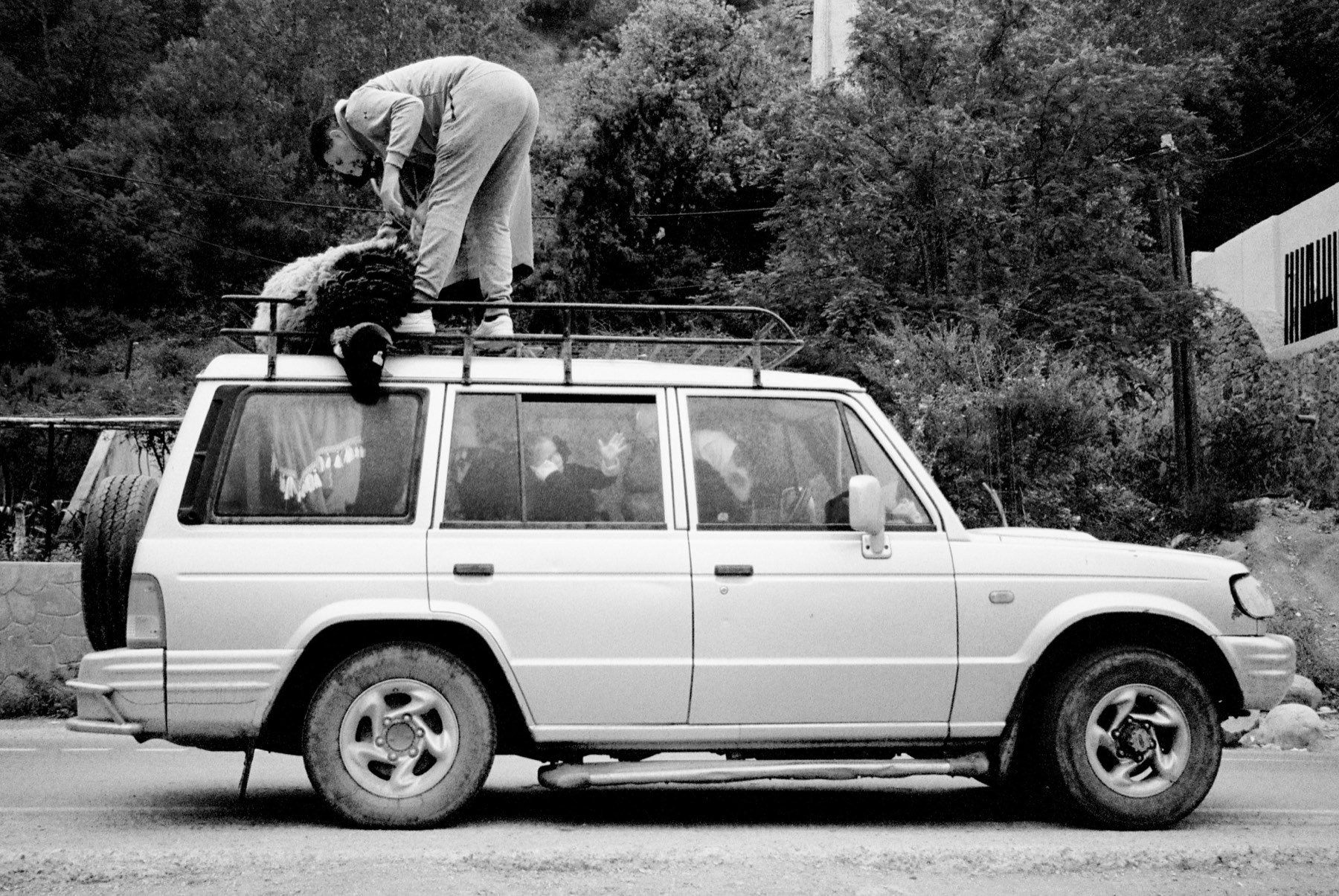 Black-and-white photograph of a person standing on a parked four-by-four's roof rack, loading gear, with passengers inside.